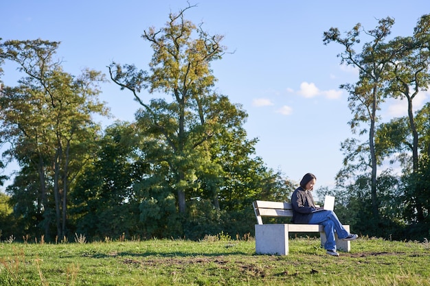 Vista lateral de una joven sentada sola en el parque en un banco usando su computadora portátil para estudiar o trabajar de forma remota f