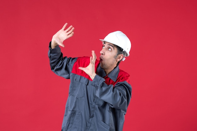 Vista lateral del joven trabajador asustado en uniforme con casco en pared roja aislada