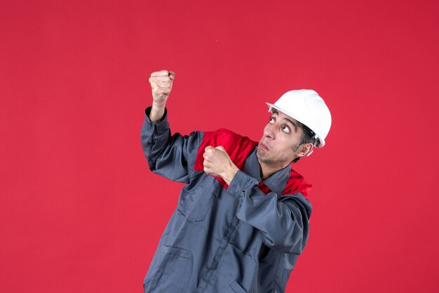 Vista lateral del joven trabajador asustado en uniforme con casco en pared roja aislada