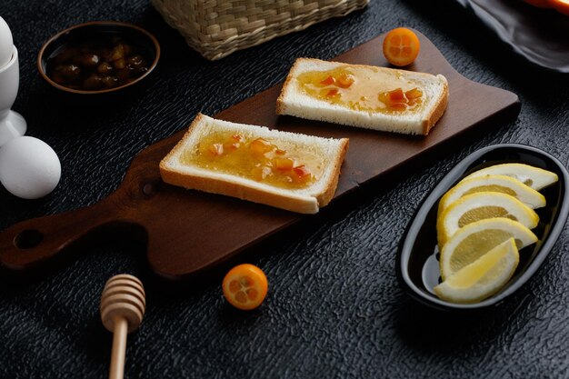 Vista lateral del juego de desayuno con mermelada untada en rebanadas de pan en la tabla de cortar y mermelada de huevos en rodajas de limón con kumquat medio cortado sobre fondo negro