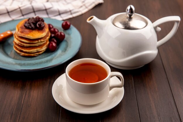 Vista lateral del juego de desayuno con panqueques y cerezas y tenedor en plato sobre tela escocesa y taza de té con tetera sobre fondo de madera