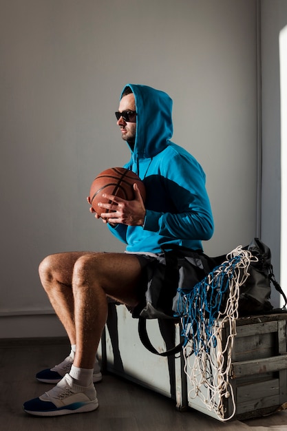 Vista lateral del jugador de baloncesto posando con capucha y gafas de sol con balón cerca del pecho