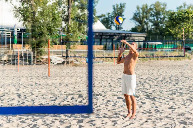 Vista lateral del jugador de voleibol masculino sin camisa practicando con pelota en la playa