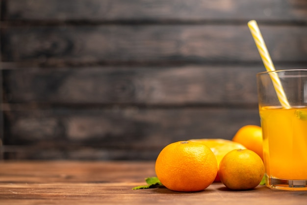 Vista lateral de jugo de naranja fresco en un vaso servido con un tubo de menta y naranjas enteras cortadas en una mesa de madera