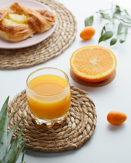 Vista lateral del jugo de naranja en salvamanteles con croissant de naranja cortado con kumquats con hojas sobre fondo blanco