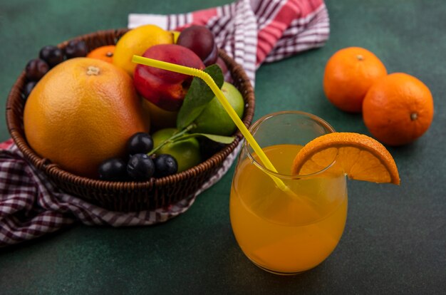 Vista lateral de jugo de naranja en un vaso con pomelo lima limón melocotón cereza ciruela naranja y ciruela en una canasta sobre un fondo verde