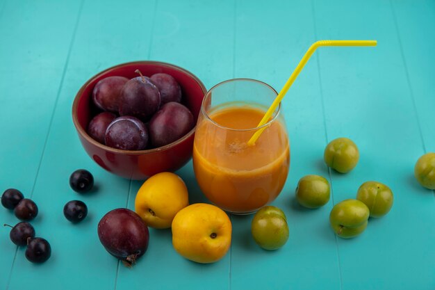 Vista lateral del jugo de nectacot con tubo para beber en vaso y tazón de pluots con ciruelas nectacots y bayas de uva sobre fondo azul.