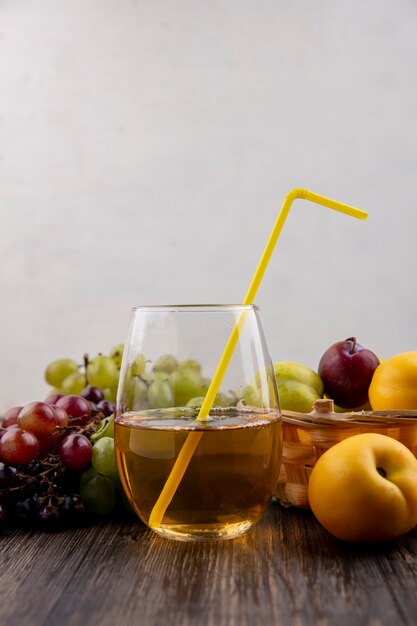 Vista lateral del jugo de uva blanca en vaso con frutas como nectacots pluots en canasta con uvas sobre superficie de madera y fondo blanco.