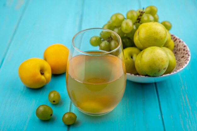 Vista lateral del jugo de uva blanca en vidrio y frutas como uva y pluots verdes en un tazón con nectacots y bayas de uva sobre fondo azul.