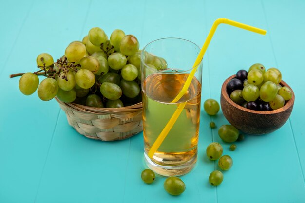 Vista lateral del jugo de uva blanca en vidrio con uvas en la canasta y en un recipiente sobre fondo azul.