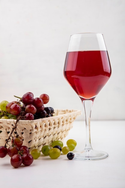 Vista lateral del jugo de uva negra en copa de vino y canasta de uvas con uvas sobre fondo blanco.