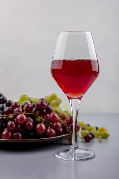 Vista lateral del jugo de uva negra en copa de vino y plato de uvas con uvas sobre superficie gris y fondo blanco.