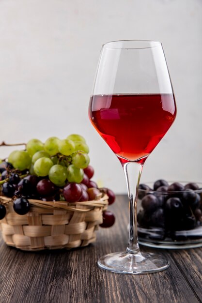 Vista lateral del jugo de uva negra en copa de vino con uvas en la canasta y en un recipiente sobre la superficie de madera y fondo blanco.