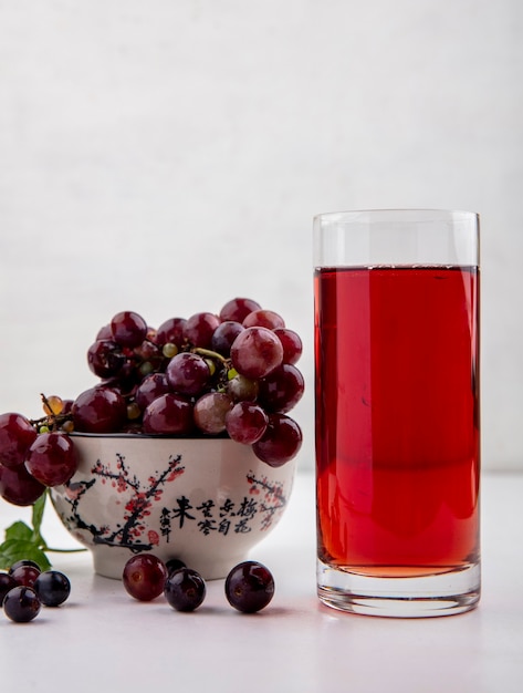 Vista lateral del jugo de uva negra en vidrio y tazón de uvas rojas sobre fondo blanco.