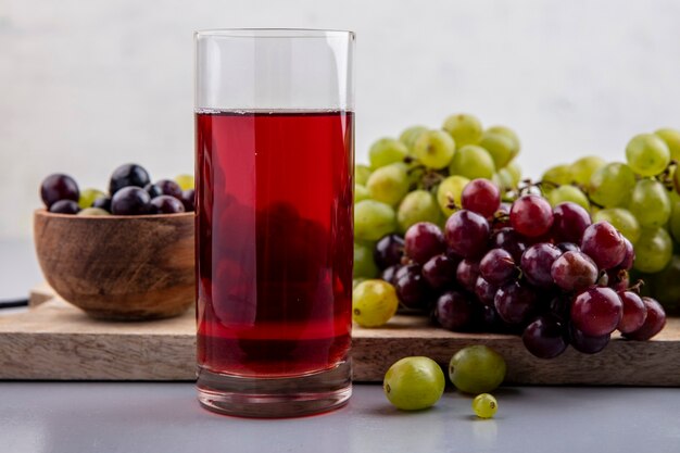 Vista lateral del jugo de uva negra en vidrio y uvas en un tazón y sobre una tabla de cortar sobre una superficie gris y fondo blanco.