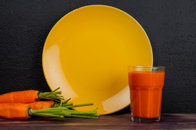 Vista lateral de jugo de zanahoria y zanahorias con plato vacío sobre superficie de madera y fondo negro