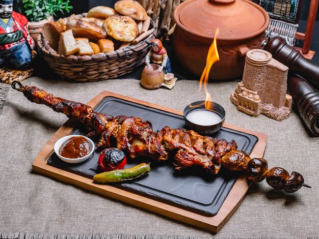 Vista lateral kebab de carne en un pincho con papas a la parrilla y verduras con salsa y fuego en un tablero
