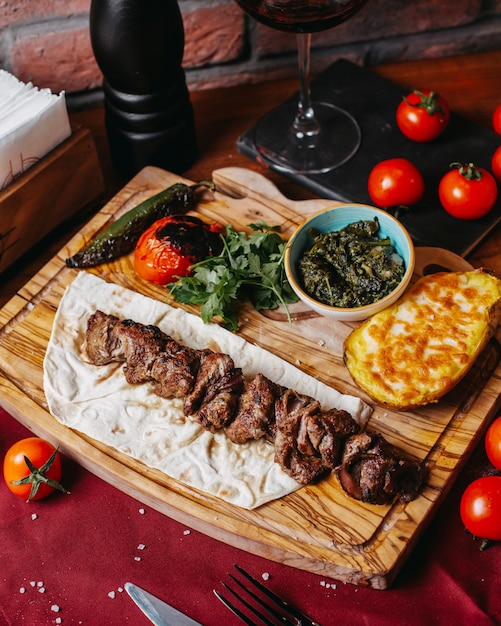 Foto gratuita vista lateral de kebab de cordero con patatas al horno y verduras sobre una plancha de madera