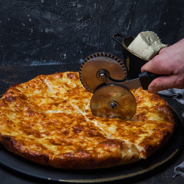 Vista lateral khachapuri con queso sulguni y cuchillo para cortar pizza y mano humana en plato oscuro