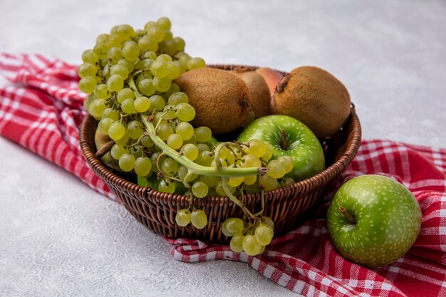 Vista lateral de kiwi con manzanas verdes, uvas y peras en una canasta sobre una toalla a cuadros rojos sobre un fondo blanco.