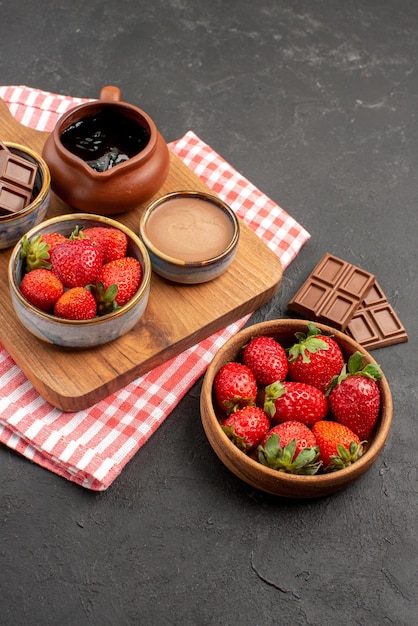 Vista lateral desde lejos fresas en tabla de cortar mantel con tazones de fresas y chocolate junto al plato de fresas en el centro de la mesa