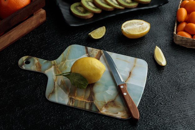 Vista lateral de limón con cuchillo en tabla de cortar con rodajas de kiwi, rodaja de lima, limón cortado y kumquats enteros sobre fondo negro