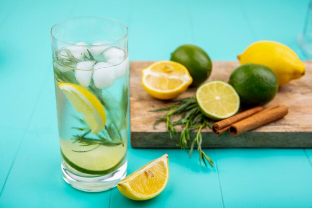 Vista lateral de limones coloridos en una tabla de cocina de madera con palitos de canela con un vaso de agua de verano en la superficie azul