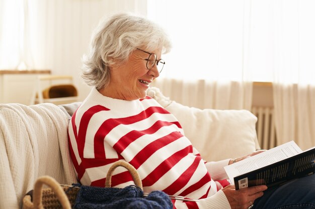 Vista lateral de la linda abuela feliz con gafas disfrutando de la lectura en el interior, sentado en el sofá con una interesante historia de detectives, sonriendo con alegría. Anciana elegante relajante en el sofá con libro