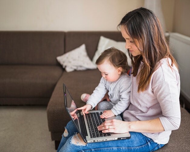 Vista lateral de la madre con su hijo sentado en el sofá usando la computadora portátil