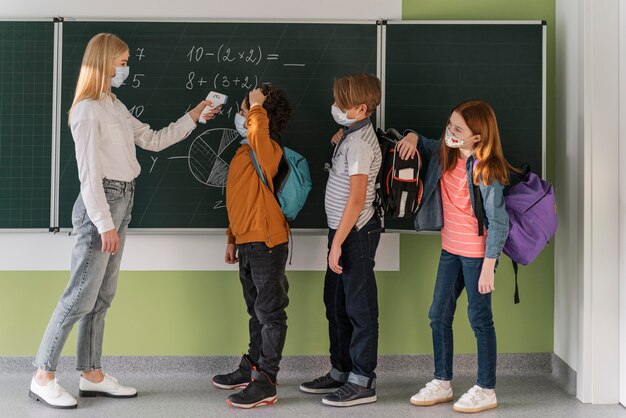 Vista lateral de la maestra con máscara médica que controla la temperatura del estudiante en la escuela