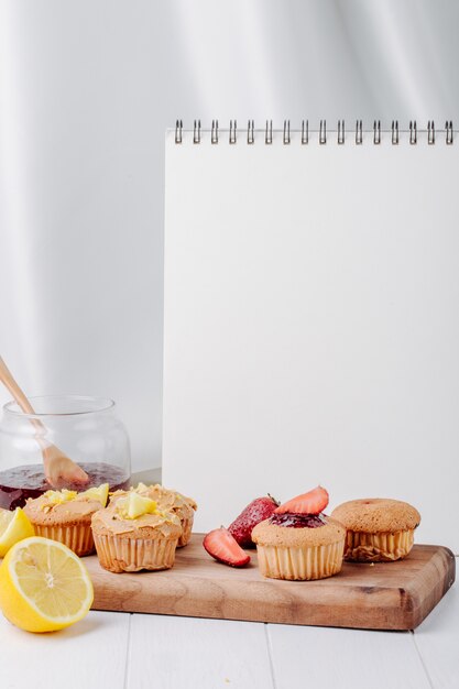 Vista lateral magdalenas con fresas y limón en una pizarra con un tarro de mermelada y un cuaderno blanco