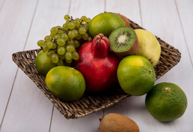 Vista lateral de mandarinas con granada, manzana, pera, uvas y kiwi en una canasta sobre una pared blanca.