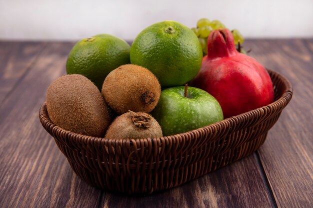 Vista lateral de mandarinas con manzanas kiwi y granada en una canasta en una pared de madera