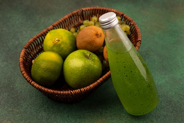 Vista lateral de mandarinas verdes con manzana, kiwi y uvas en una canasta con una botella de jugo en una pared verde