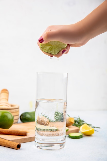 Foto gratuita vista lateral de la mano femenina exprimiendo limón fresco en un vaso de agua sobre una superficie blanca