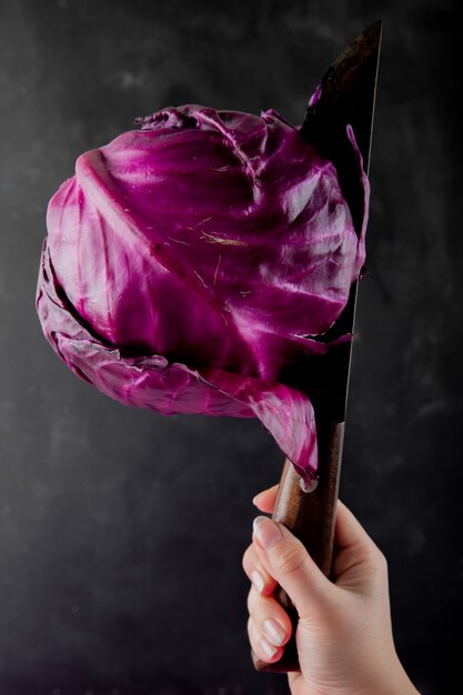 Vista lateral de la mano de mujer con cuchillo cortando repollo morado sobre fondo negro con espacio de copia