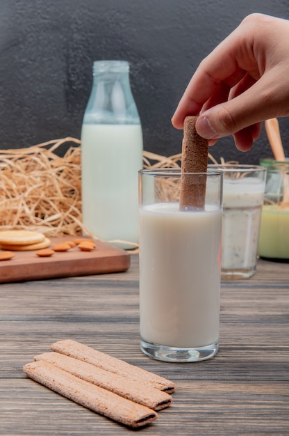 Vista lateral de la mano que sostiene la galleta con un vaso de leche y galletas de almendras en la tabla de cortar sopa de yogurt leche condensada en superficie de madera y superficie negra