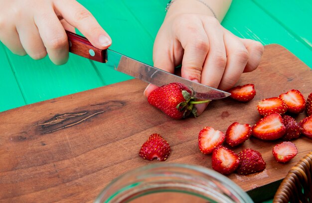 Vista lateral de manos cortando fresas con cuchillo en la tabla de cortar en superficie verde