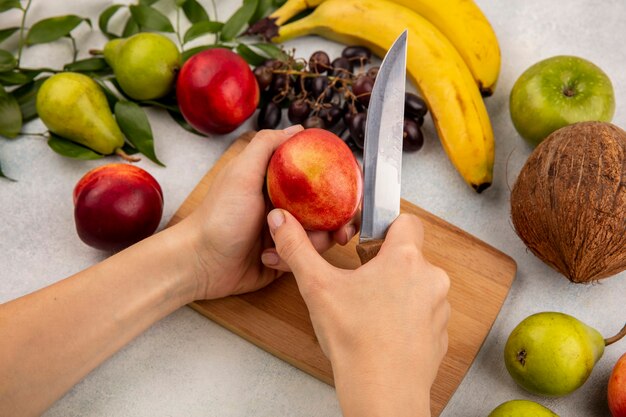 Vista lateral de las manos cortando melocotón con un cuchillo en la tabla de cortar y manzana de plátano coco pera uva con hojas sobre fondo blanco.