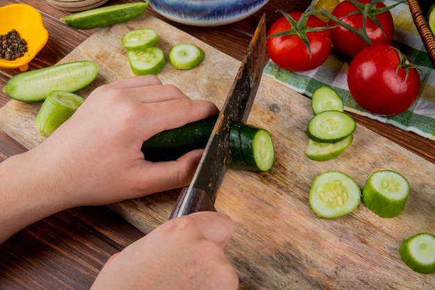 Vista lateral de manos cortando pepino con cuchillo en la tabla de cortar con tomates pimienta negra sobre superficie de madera