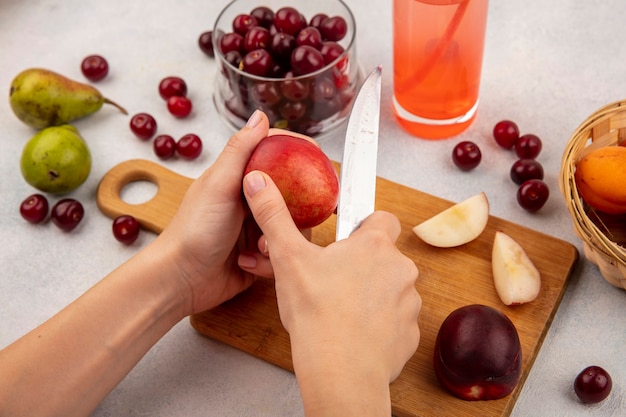 Vista lateral de las manos femeninas cortando melocotón con un cuchillo en la tabla de cortar y jugo de cereza con tarro de cereza y canasta de albaricoque con peras sobre fondo blanco.