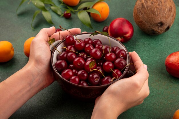 Vista lateral de las manos femeninas sosteniendo un tazón de cerezas con patrón de duraznos, albaricoques, cerezas, coco y hojas sobre fondo verde