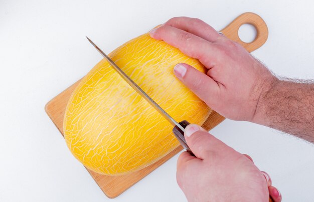 Vista lateral de manos masculinas cortando melón con cuchillo en la tabla de cortar sobre fondo blanco.