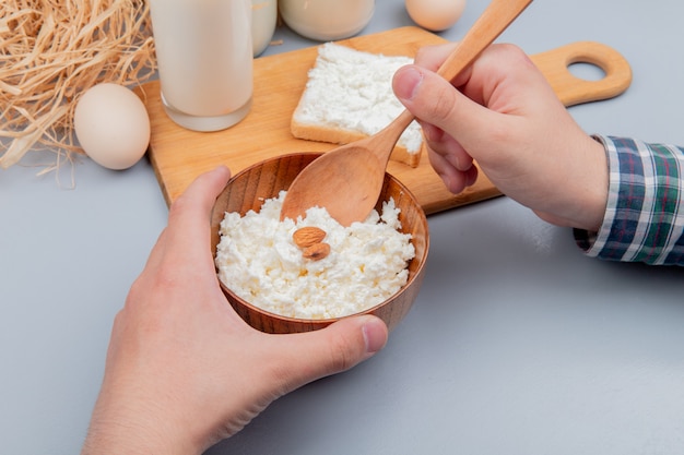 Vista lateral de manos masculinas que sostienen un tazón de requesón y una cuchara de madera con una rebanada de pan untada con leche de requesón en la tabla de cortar y huevos de paja en la superficie azul