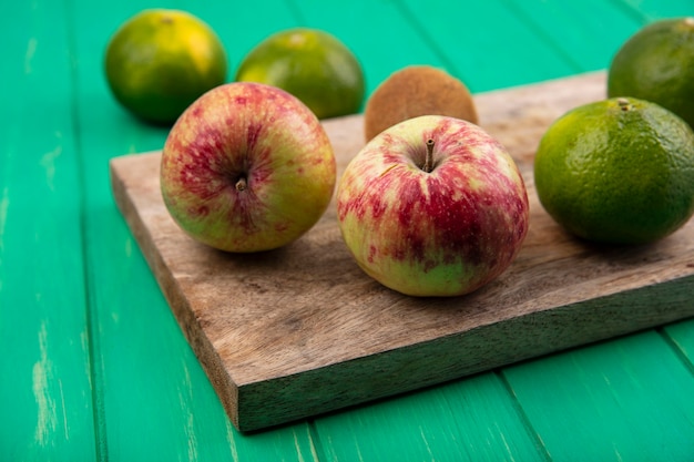 Vista lateral de manzanas con mandarinas sobre una tabla para cortar en una pared verde