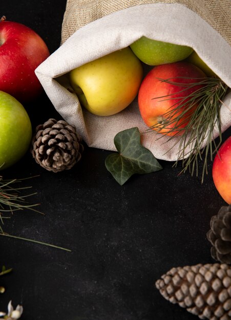 Vista lateral de manzanas multicolores en una bolsa de arpillera con conos de abeto sobre una mesa negra