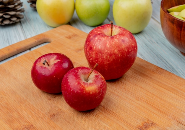 Vista lateral de manzanas rojas en tabla de cortar con las amarillas y verdes sobre fondo de madera