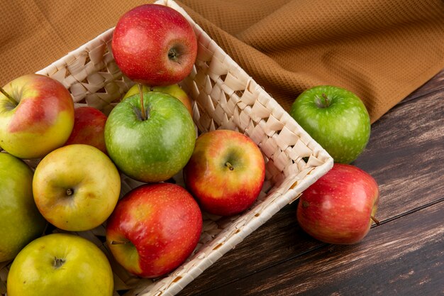Vista lateral de manzanas verdes y rojas en una cesta sobre una toalla marrón sobre un fondo de madera