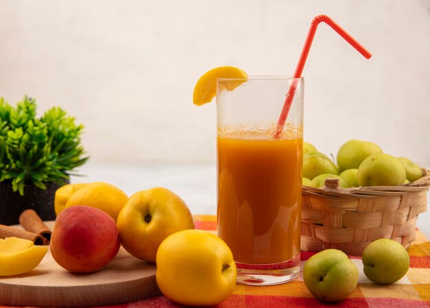 Vista lateral de melocotones amarillos dulces en una tabla de cocina de madera con melocotón rosado anaranjado con jugo de melocotón con ciruelas cereza verde en un cubo sobre un mantel a cuadros sobre un fondo blanco