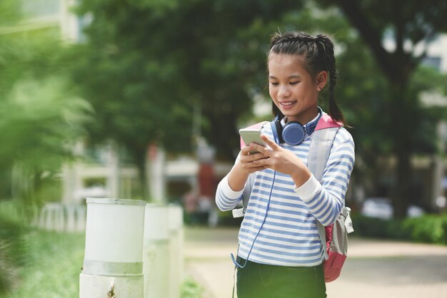 Vista lateral del mensaje de texto de una niña asiática a sus padres después de la escuela de pie al aire libre en un agradable día de primavera